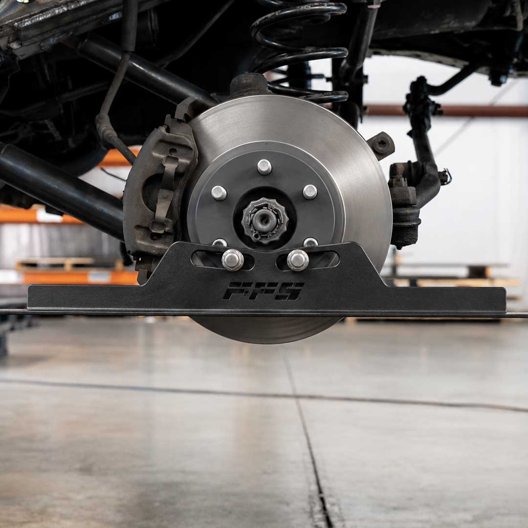 A close-up of a black PPS brake shield mounted in front of a vehicle's brake rotor, with the suspension and wheel assembly visible in a garage setting, using Far From stocks Toe Alignment Tool.