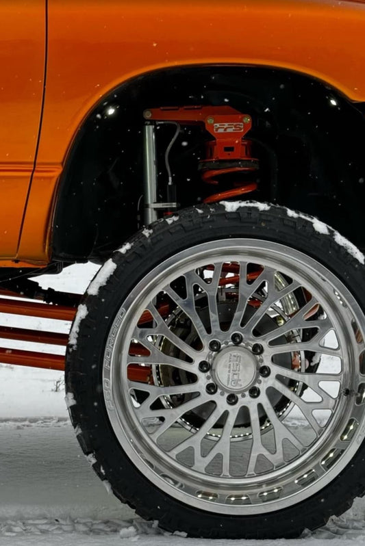 A close-up of a vehicle's front coil leveling kit on a snow-covered ground, showcasing the wheel and the upgraded suspension parts.