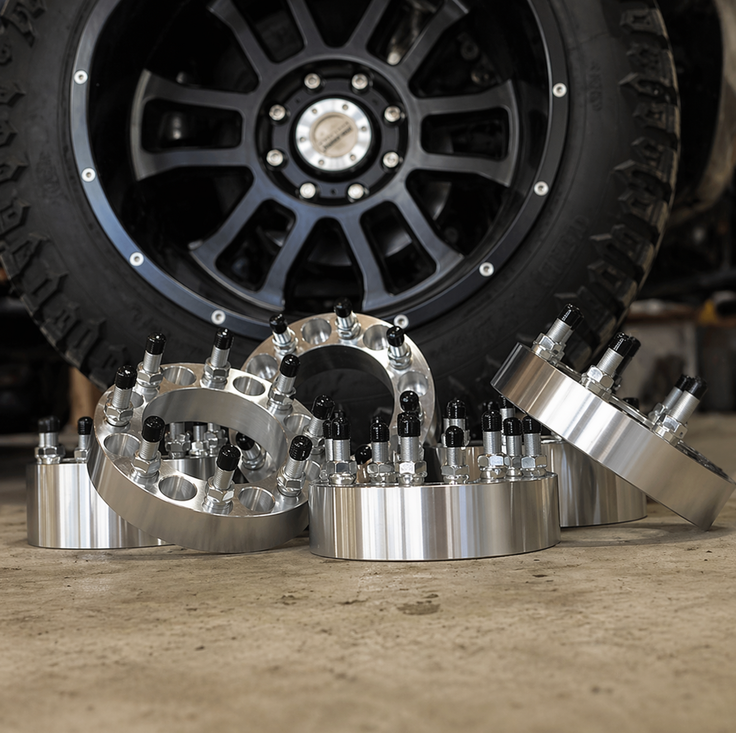 Billet aluminum 8-lug truck wheel spacers with studs displayed in front of an aftermarket truck wheel in a shop setting.